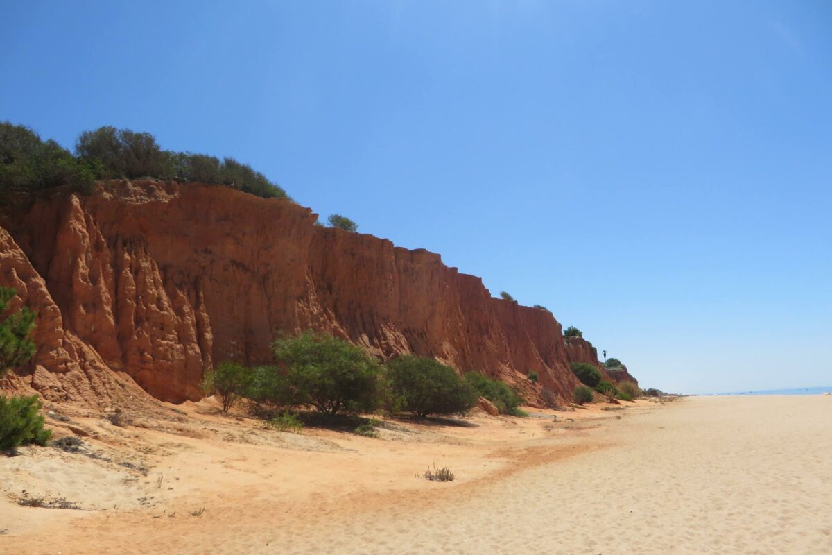 Dark red cliff edge overlooking a beautiful beach on a sunny day in the Algarve, Portugal.