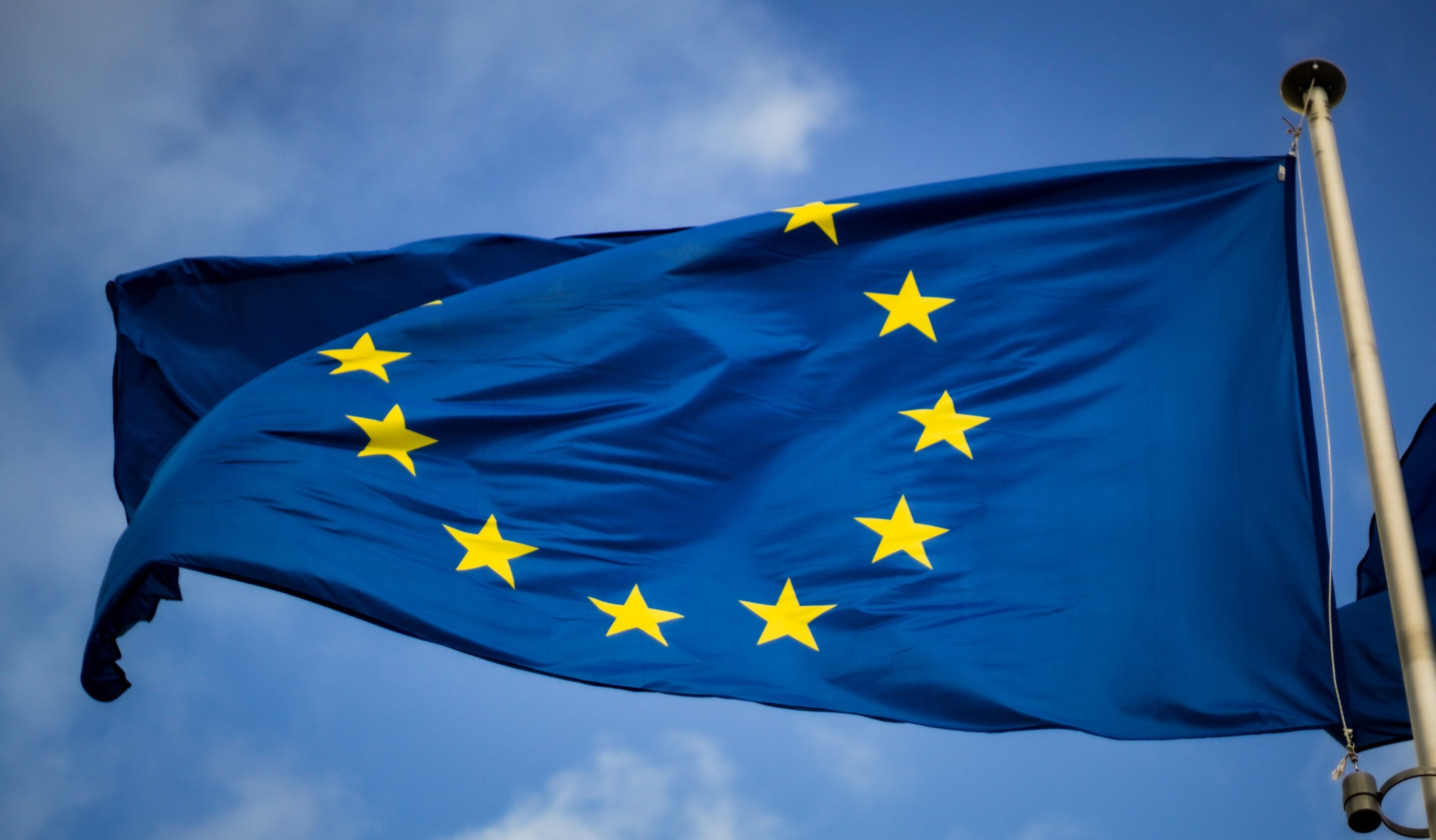 The EU flag, blue with yellow stars, is seen blowing in the wind on a sunny day with blue sky in the background.