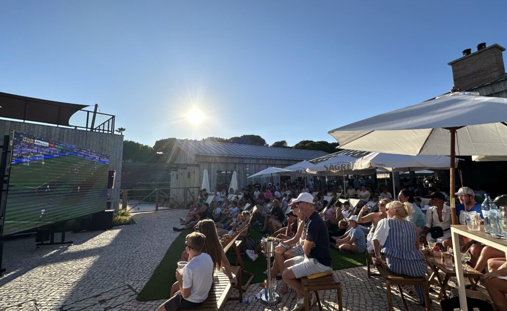 Crowd of people watching a football match outside on a sunny day.